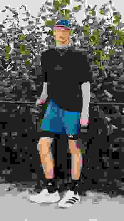 A person wearing a black shirt, blue shorts, and sneakers stands in front of a lush, green foliage background.