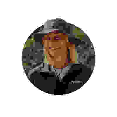 A smiling person wearing a Terrex jacket and a wide-brimmed hat stands in a natural, outdoor setting with greenery in the background.
