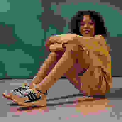 A young woman with curly hair sits on the floor, wearing a yellow top and beige pants, with sneakers on her feet, against a blurred teal background.
