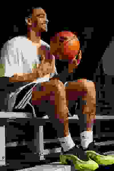 A smiling young man with tattoos holds a basketball while sitting on a bench in a dimly lit setting.
