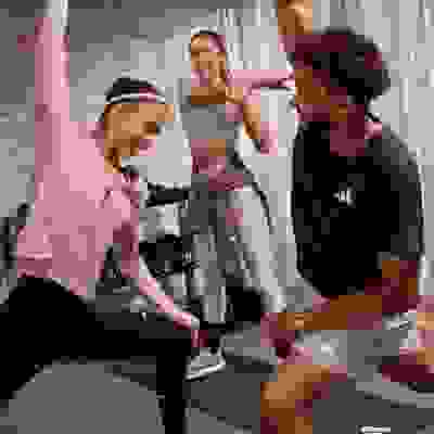 Three individuals, two women and one man, are engaged in a fitness activity in a gym setting with a brick wall backdrop.