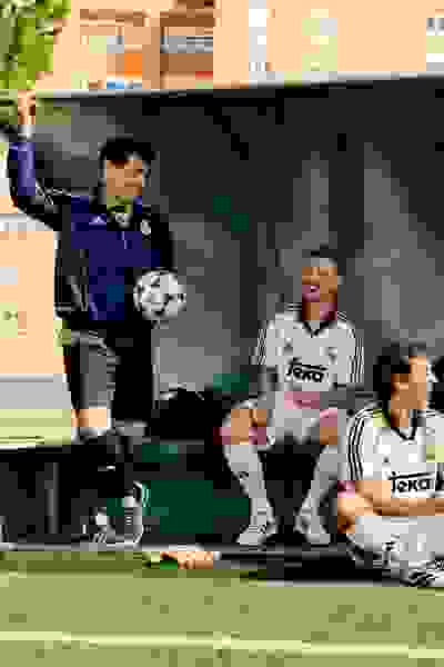 The image shows a group of soccer players on a field, with one player holding a soccer ball and another player sitting on the bench, while a building can be seen in the background.