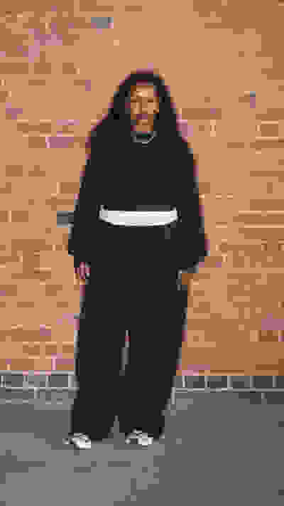 A person wearing a black hooded sweatshirt and loose-fitting pants stands in front of a brick wall.