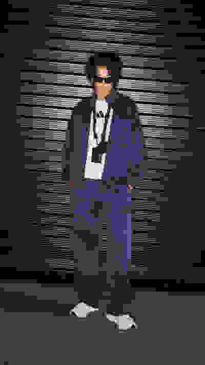 A person wearing a purple outfit, including a jacket and pants, stands in front of a dark, striped background.