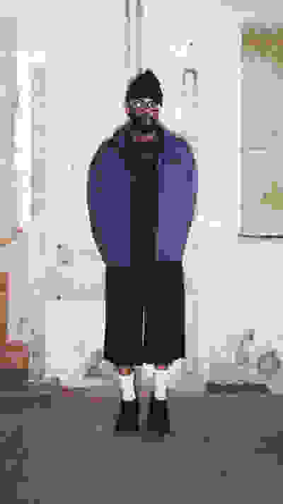 A person wearing a purple jacket and black pants stands in front of a dilapidated brick wall.