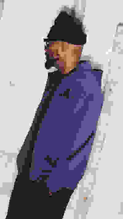 The image shows a person wearing a purple jacket and a black hat, standing in front of a white wall.