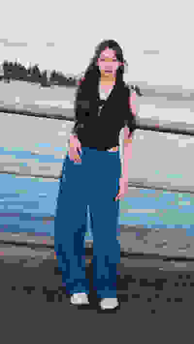 The image shows a young woman with long dark hair wearing a black top and blue pants, standing on a wooden dock with a body of water and mountains in the background.