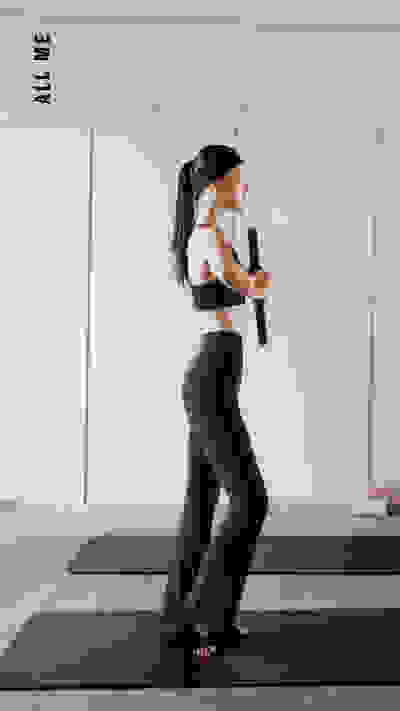 Eine Frau in Sportbekleidung steht in einem minimalistischen Studio mit weißen Wänden auf einer Yogamatte und hält ein Widerstandsband in der Hand.