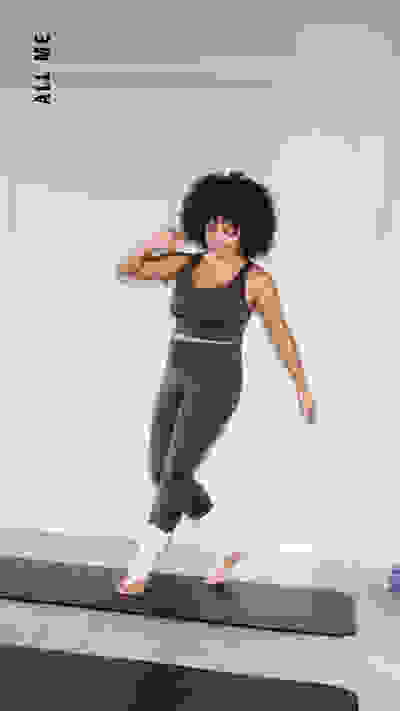 Eine Frau mit lockigem Haar steht in einem hellen, minimalistischen Raum auf einer Yogamatte.