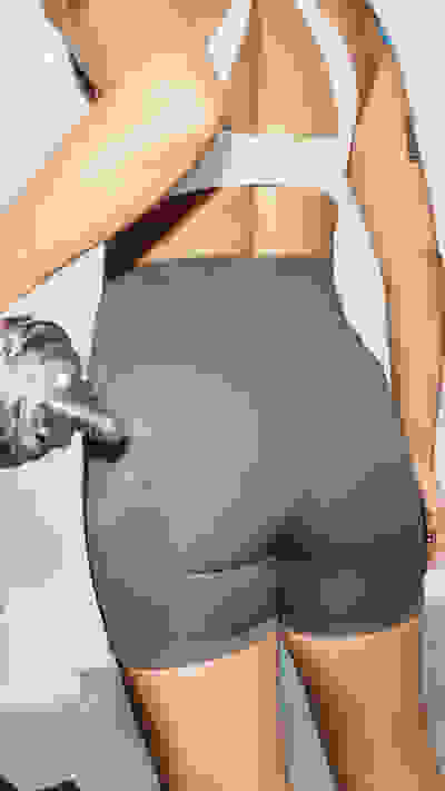 Das Bild zeigt eine Person im unteren Rumpfbereich in eng anliegenden grauen sportlichen Shorts, die eine Hantel in den Händen hält, vor einem schlichten weißen Hintergrund.