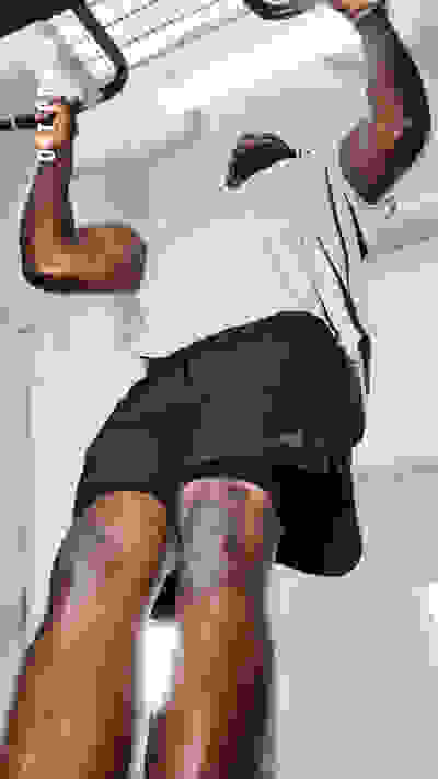 Das Bild zeigt eine Person, die eine Klimmzugübung auf einem Fitnessgerät durchführt. Im Hintergrund sind eine weiße Wand und ein Metallrahmen zu sehen.