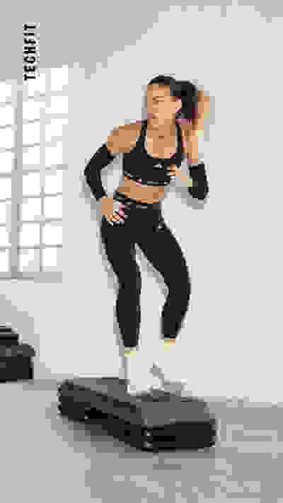 Eine Frau in Sportbekleidung springt in einem hellen, offenen Raum mit großen Fenstern auf eine schwarze Übungsplattform.