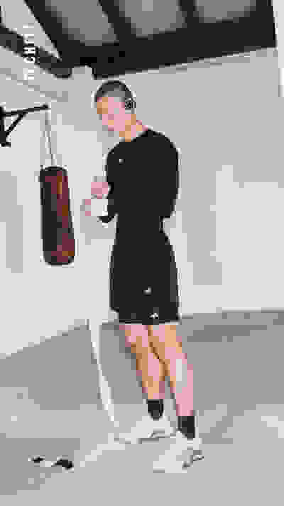 Eine Person in schwarzer Sportbekleidung steht in einem gymähnlichen Umfeld, hält eine Boxtasche in der Hand und trägt Kopfhörer.