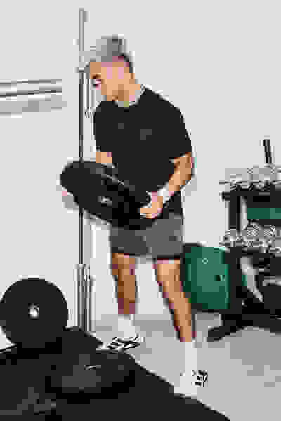 Eine Person in einem schwarzen Hemd und schwarzen Shorts hebt eine schwere Langhantel in einem Fitnessstudio, das von verschiedenen Trainingsgeräten umgeben ist.