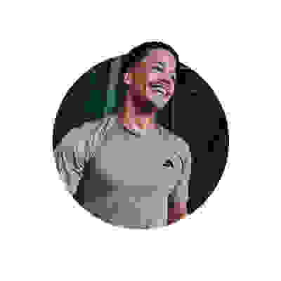 A smiling young man wearing a beige Adidas shirt stands in front of a dark green background.