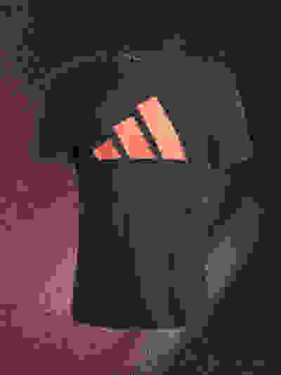 A black t-shirt with the Adidas logo prominently displayed in a bright coral color against a dark, textured background.