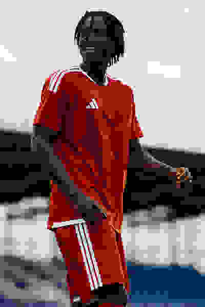 A smiling individual wearing a red Adidas sports jersey stands in front of a body of water and a cloudy sky.