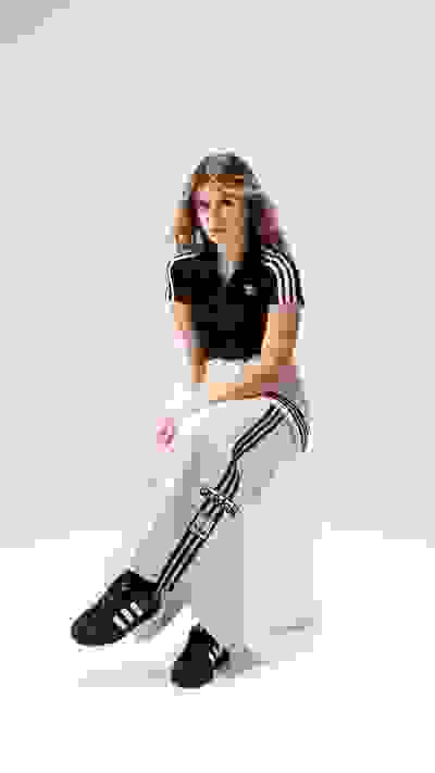 A young woman wearing a black Adidas top and beige pants with black stripes sits on a white background.