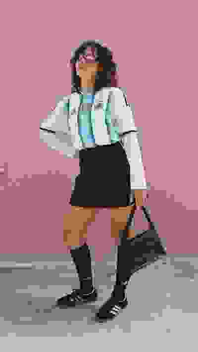 A young person wearing an Argentina national team jersey, black shorts, and black socks stands in front of a pink wall, holding a black bag.