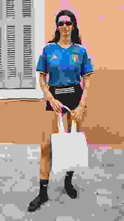A person wearing a blue sports jersey, black shorts, and carrying a white tote bag stands in front of a pink-colored building with shuttered windows.