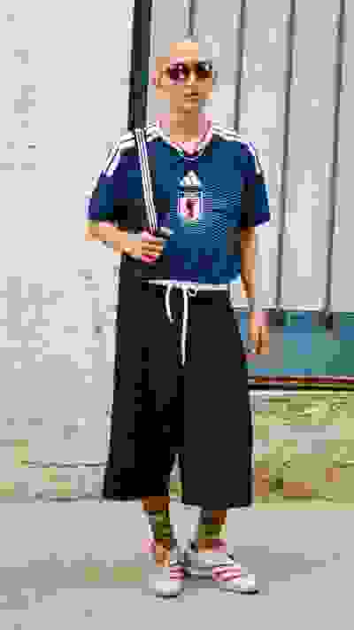 A person wearing a blue and white sports jersey, black shorts, and red and white sneakers stands in front of a weathered, blue-painted wall.