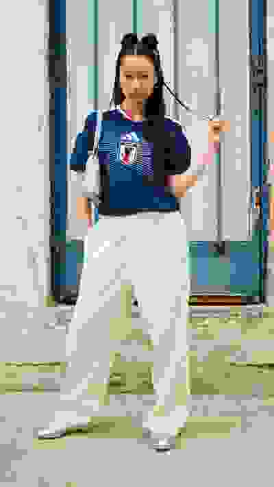The image shows a young woman wearing a blue jersey and white pants, standing in front of a blue and white building.