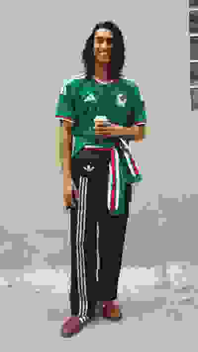 A smiling individual wearing a green jersey and black pants stands in a plain indoor setting.