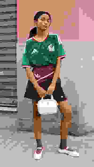 The image shows a young woman wearing a green and white soccer jersey, standing in front of a pink wall with a metal shutter in the background. She is holding a white handbag and appears to be posing for the photograph.
