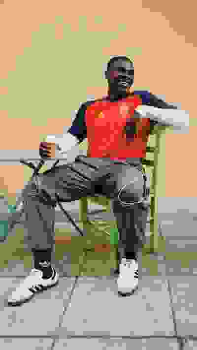 A man in a red and blue sports jersey is sitting on a green chair, holding a cup and appearing to be relaxed.