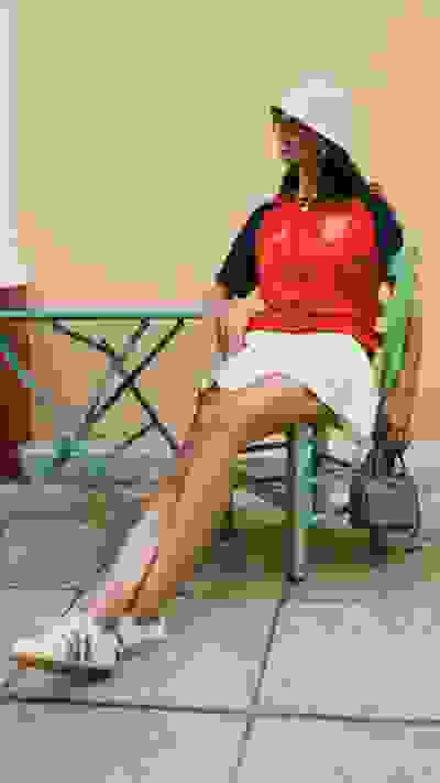 A woman in a red top and white skirt is sitting on a colorful chair, with a beige wall in the background.