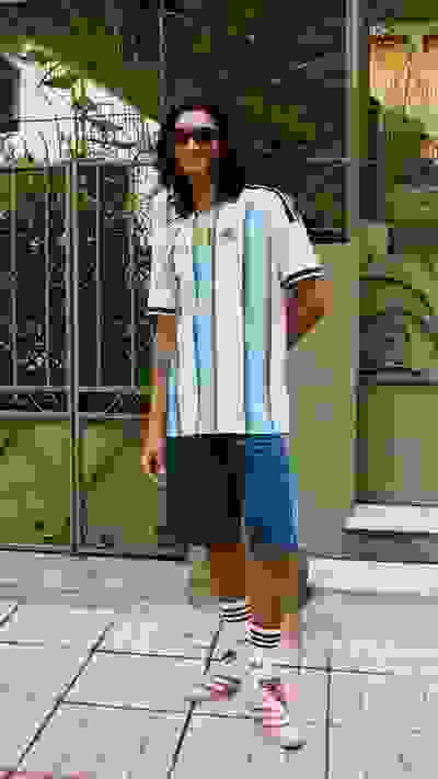 A person wearing a light blue and white jersey, blue shorts, and white sneakers stands in front of a decorative metal gate with a lush, green background.