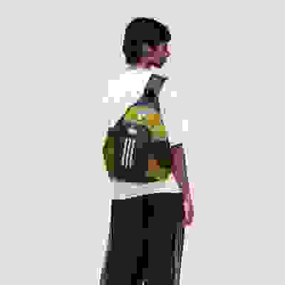 A young person wearing a white t-shirt and black pants, carrying a yellow and black backpack, stands against a plain light-colored background.