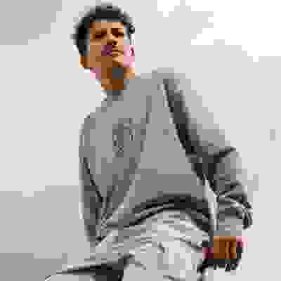 A young man with curly hair wearing a light blue sweatshirt stands against a cloudy sky background.