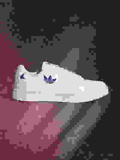 A white Adidas sneaker with blue logo sits on a red and black textured background, creating a striking visual contrast.