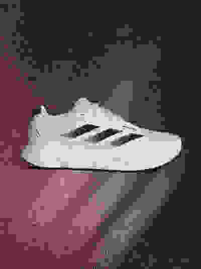 A white and black athletic shoe is prominently displayed against a dark, textured background with a reddish-pink gradient.