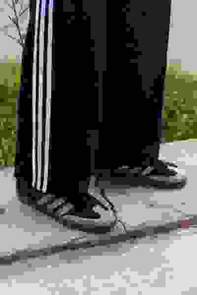 De afbeelding toont een zwarte broek met witte strepen aan de zijkanten en een paar grijze en witte sneakers op een betonnen oppervlak met een grasachtige achtergrond.