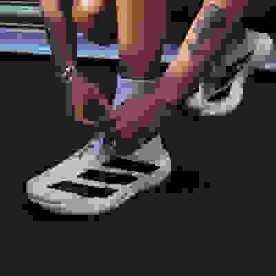 The image shows a person's lower body and feet, with the person wearing white sneakers and a tattoo visible on their arm.