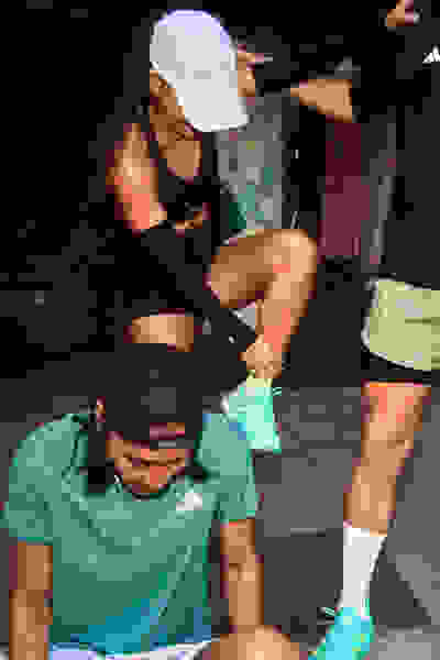 The image shows a person in a green athletic shirt and another person in black athletic clothing assisting them on what appears to be a fitness or training facility.