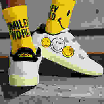 The image shows a pair of white Adidas sneakers with yellow smiley face designs and the text "Smile World" printed on the socks.