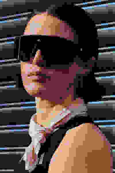 Una persona che indossa occhiali da sole e una sciarpa sta di fronte a uno sfondo in metallo con l'otturatore.