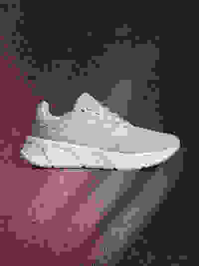 A white and gray athletic shoe is displayed against a dark red and black background, with a subtle lighting effect creating a sense of depth and drama.