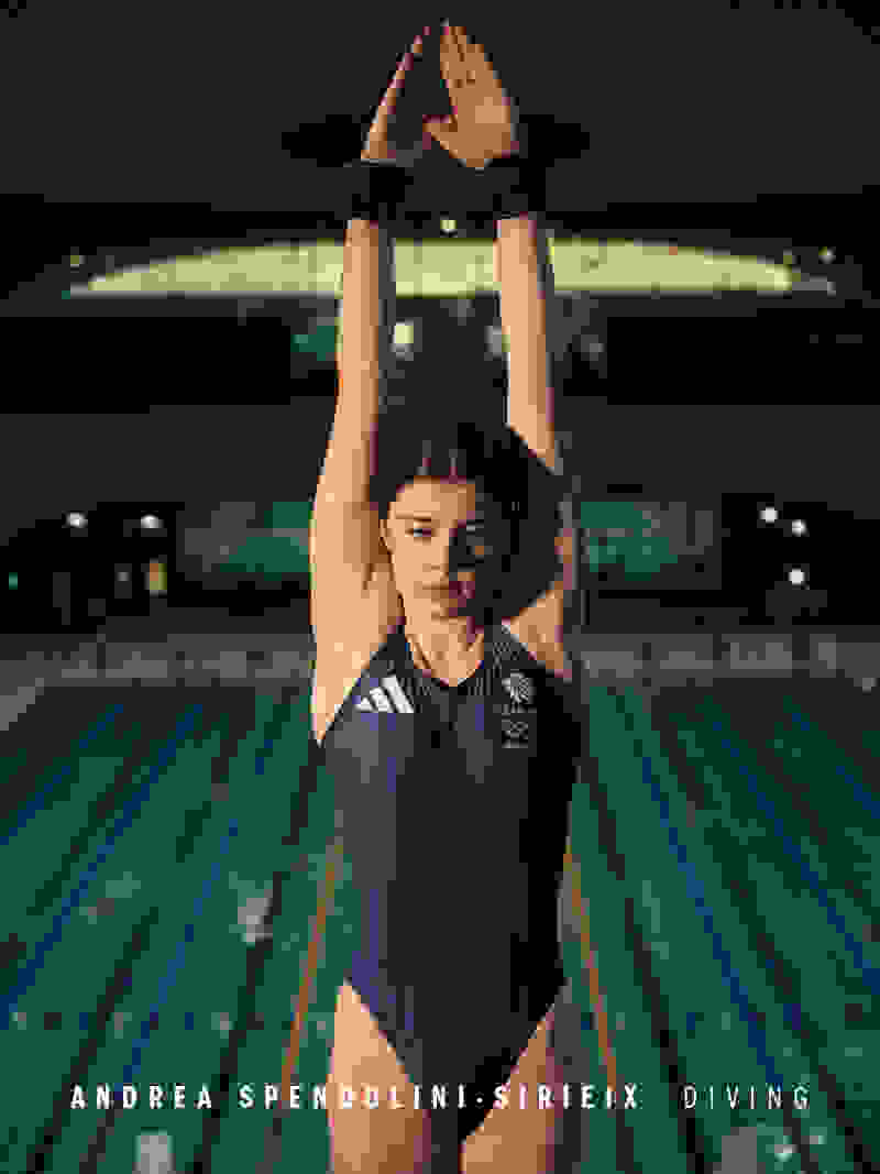 The image shows a woman in a diving suit standing in front of a swimming pool, with her arms raised overhead in a diving pose, against a backdrop of stadium lights and a nighttime sky.