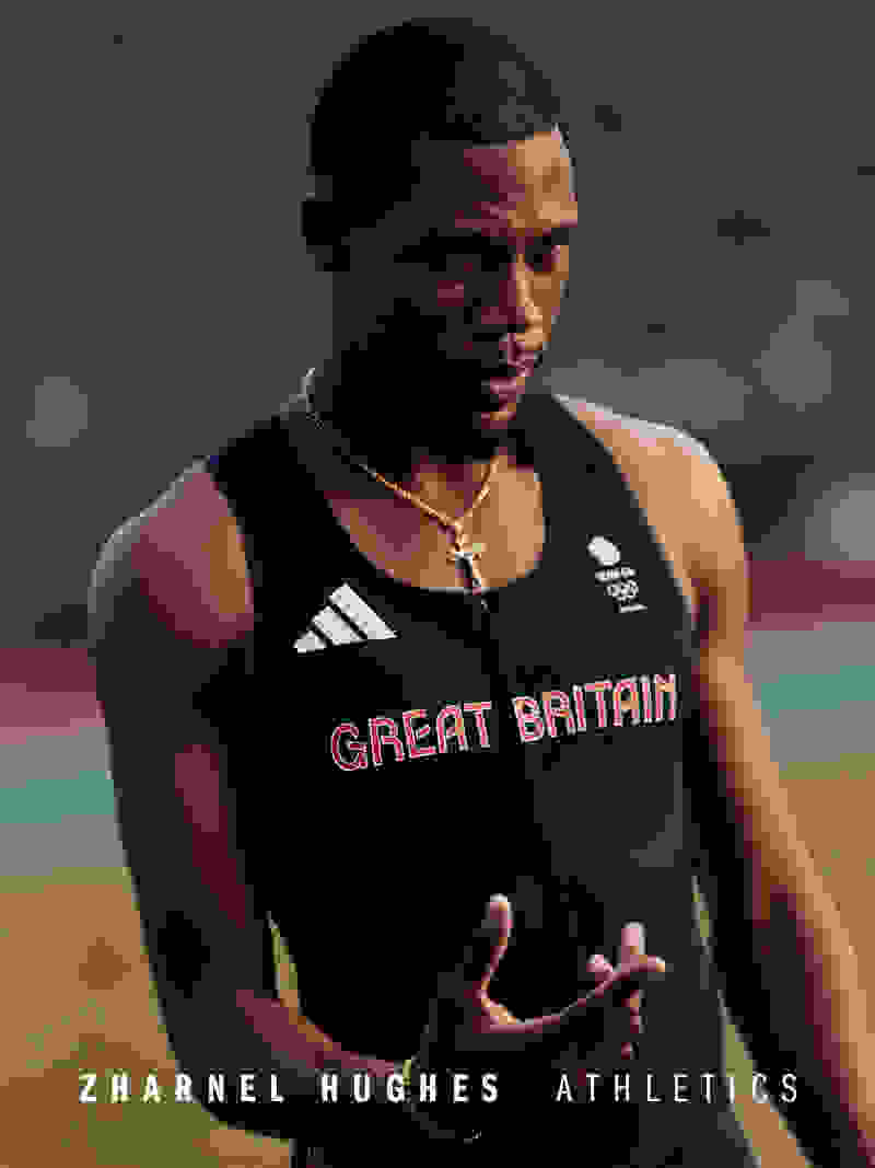 The image shows a muscular, shirtless Black man wearing a Great Britain jersey and standing in an outdoor athletic setting, with a focused expression on his face.