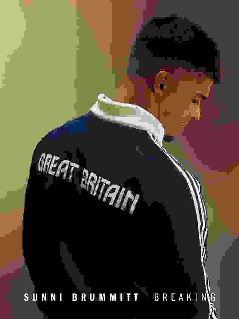 The image shows a person wearing a navy blue jacket with the text "GREAT BRITAIN" printed on the back, standing in a dimly lit setting.