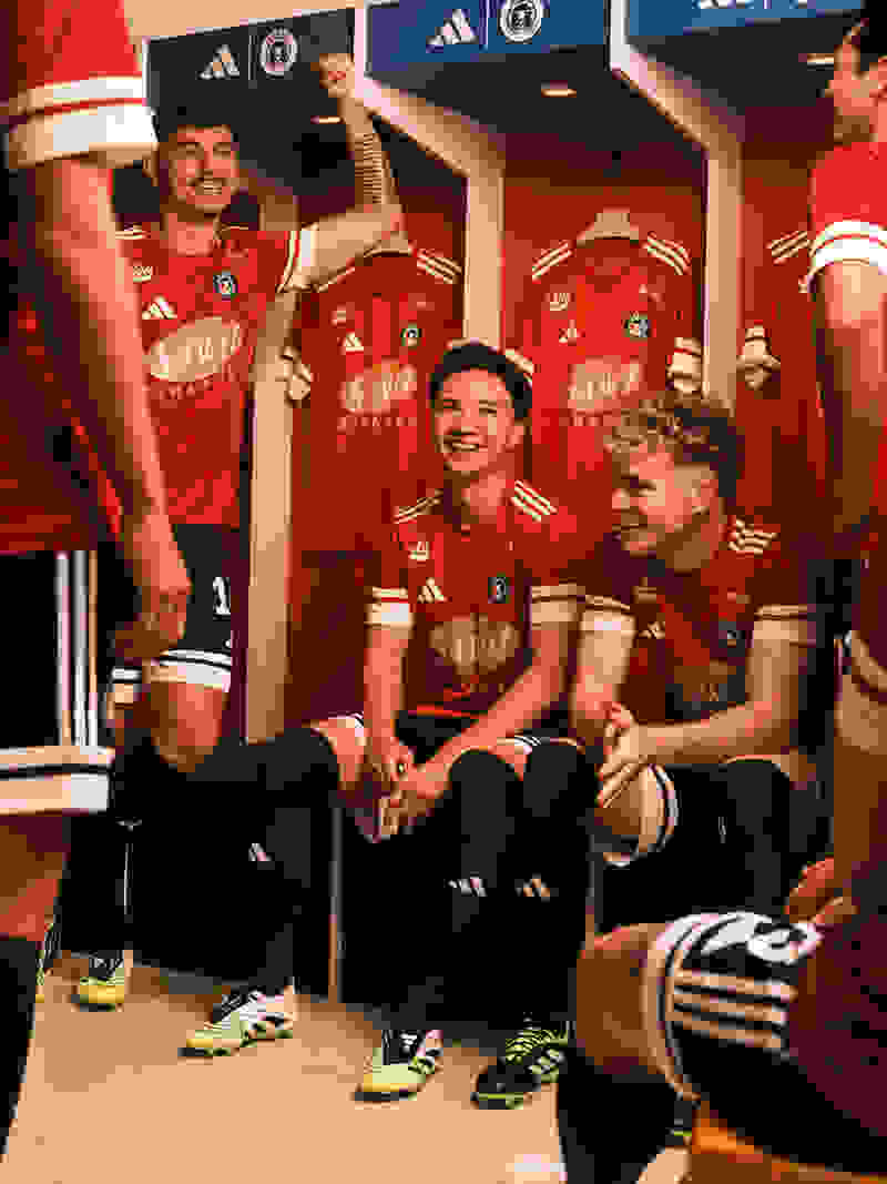 The image shows a group of young soccer players sitting in a locker room, wearing red and white uniforms and smiling at the camera. The background features shelves with additional uniforms and sports equipment.