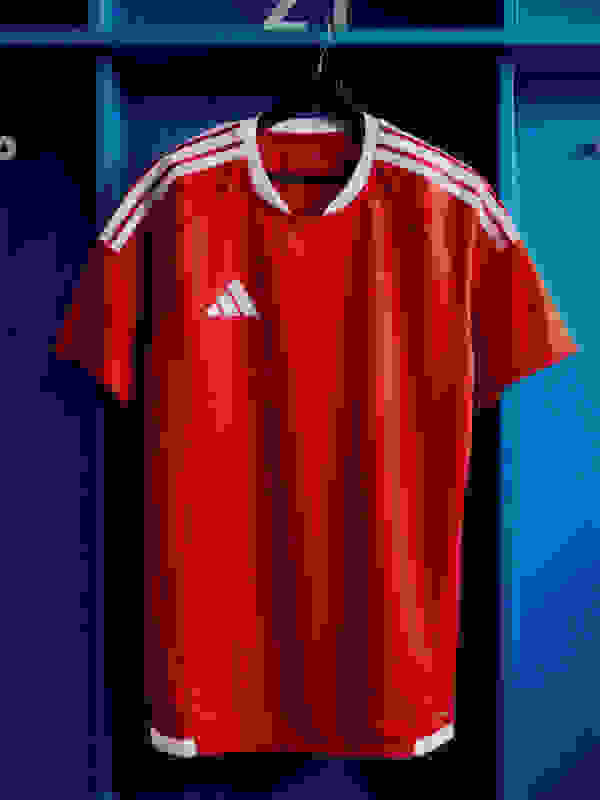 A vibrant red Adidas soccer jersey hangs on a blue locker, with white stripes and the Adidas logo prominently displayed.