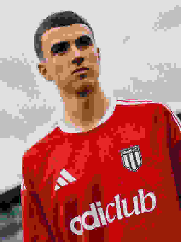 A young man wearing a red soccer jersey with the "Adiclub" logo stands in an outdoor setting with a cloudy sky in the background.