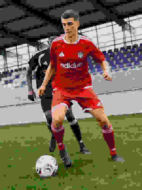 A young soccer player in a red jersey is dribbling a ball on a grassy field, with a stadium-like structure visible in the background.