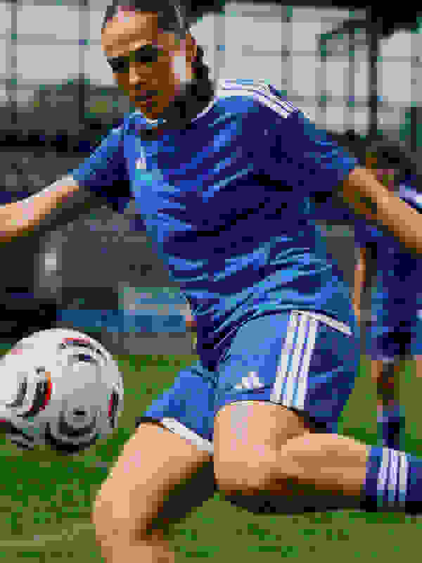 A female soccer player in a blue uniform is dribbling a soccer ball on a grassy field, with a sports facility visible in the background.