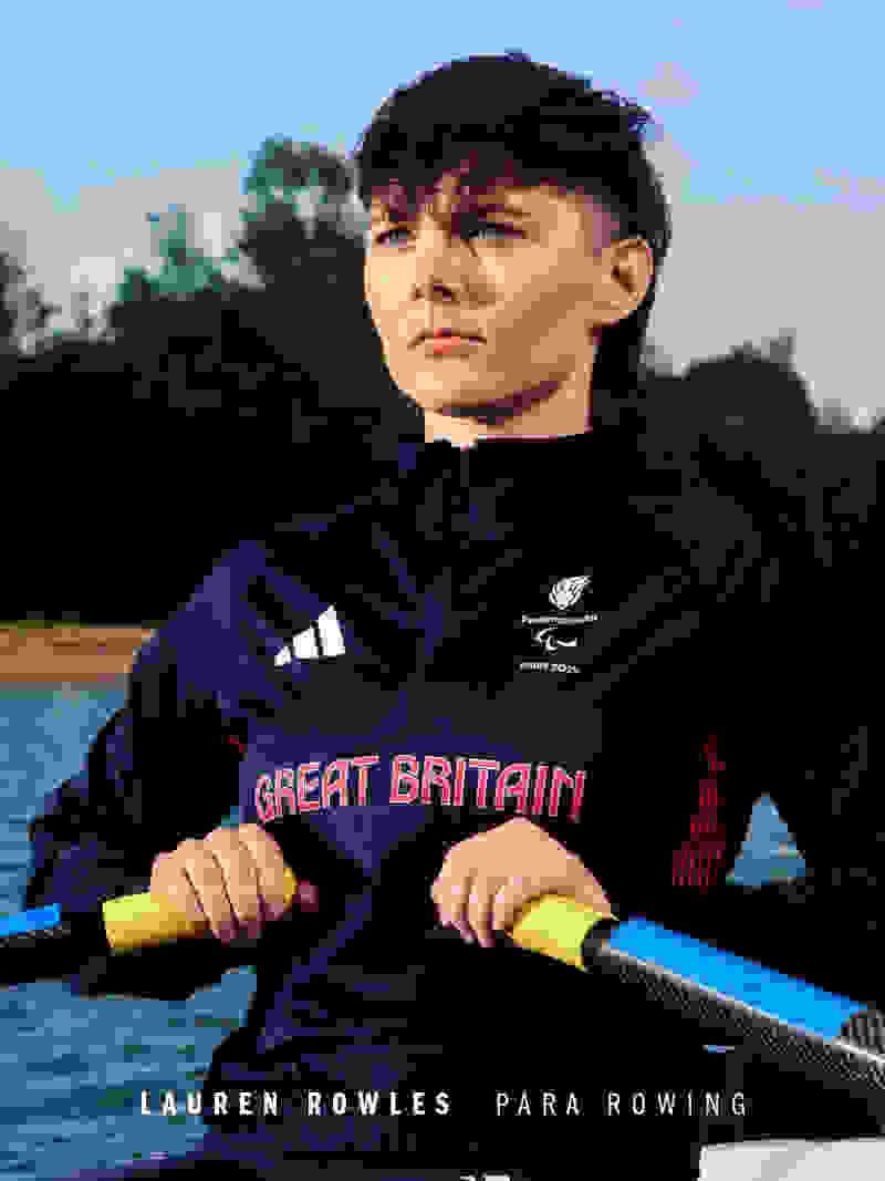 A young person wearing a navy blue athletic jacket with "Great Britain" printed on it, holding a rowing oar and standing in front of a body of water with trees in the background.